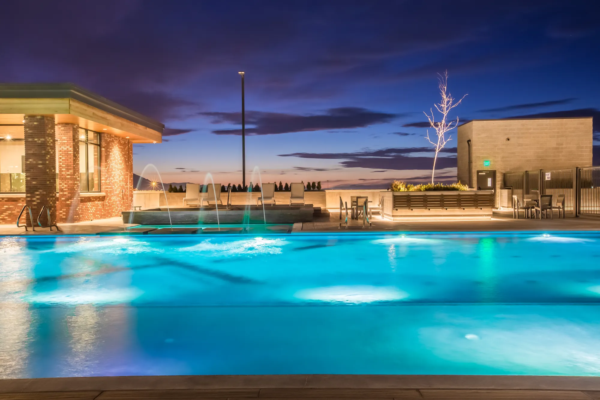 The 4th West rooftop pool lit and glowing at night, fountains arcing over the water, skyline beyond